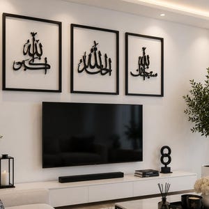 May include: Three framed black wall art pieces with Arabic calligraphy hang above a large flat-screen TV. The room features a white entertainment center, a decorative plant, and a modern aesthetic.