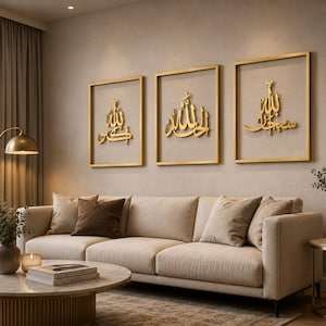May include: Three framed gold-colored Islamic calligraphy art pieces hang above a beige sofa. The artwork features ornate Arabic script within gold frames. The room has a neutral color palette with a gold floor lamp and a marble coffee table.