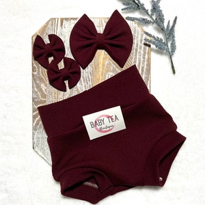 May include: Burgundy baby bloomers with a textured fabric and a small tag that reads "BABY TEA Boutique". The image also includes three burgundy fabric hair bows.