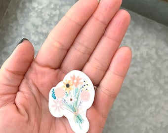 May include: A hand holding a white sticker with a colorful floral bouquet design. The bouquet features pink, yellow, and blue flowers with green stems and leaves.