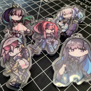 May include: A collection of five anime-style acrylic keychains. Each keychain features a unique character design with vibrant colors, including pink, blue, and green. The characters are depicted in various poses, some holding weapons.