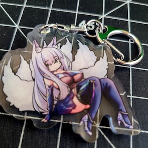 May include: A clear acrylic keychain featuring a cartoon anime character with long lavender hair and nine white fluffy tails. The character is wearing a purple and blue outfit. The keychain has a silver clasp and a green accent.