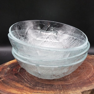 May include: A stack of three clear glass bowls featuring a textured floral pattern on the exterior. The bowls have a smooth interior surface and a rounded rim, with the embossed flower design visible through the transparent glass material.