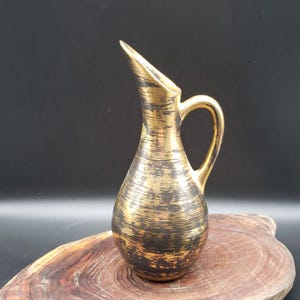 May include: A vintage gold and black ceramic pitcher with a handle and a slender spout. The pitcher has a textured, brushed design and sits on a wooden surface. The overall aesthetic is retro and decorative.
