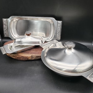 May include: A collection of vintage silver serving dishes. The set includes a large rectangular tray, a covered oval dish, and a smaller rectangular dish, all with decorative handles. The covered dish has a wooden knob on the lid.
