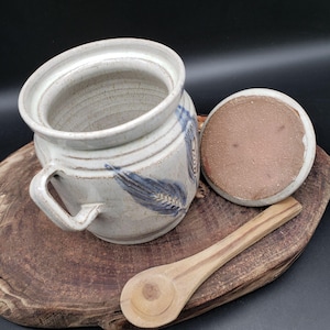 May include: A handmade, off-white ceramic butter dish with a lid and a handle. The dish features blue wheat stalk designs. A wooden spoon rests beside the dish on a wooden cutting board. The lid is off to the side.