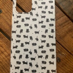 May include: A white phone case with a black cat pattern. The case has a cutout for the camera lens.