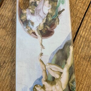 Puede incluir: Una funda transparente para tel&eacute;fono con una imagen impresa de La Creaci&oacute;n de Ad&aacute;n de Miguel &Aacute;ngel. La imagen muestra a Dios tendi&eacute;ndole la mano a Ad&aacute;n, que est&aacute; acostado sobre una roca.