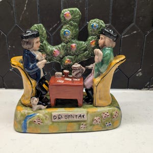 May include: A ceramic figurine depicting two figures seated at a table, playing cards. The figures are dressed in period clothing, with a green tree-like structure behind them. The base is decorated with flowers and the text "DR. SYNTAX".