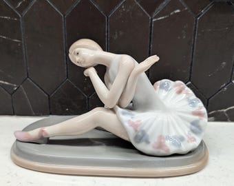 以下が含まれることがあります： 手の上に頭を乗せて座っているバレリーナの磁器製フィギュア。彼女はピンクとブルーの花柄のアクセントがついた白いチュチュを着ています。フィギュアは灰色の台座の上に座っています。