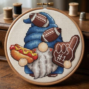 May include: Cross-stitch embroidery of a gnome in a blue outfit, holding two footballs, a hot dog, and a foam finger. The gnome has a white beard and is set in a wooden embroidery hoop. The foam finger says #1.