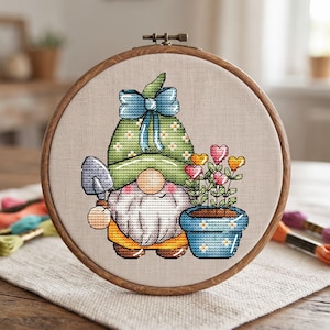 May include: A cross-stitch embroidery in a round wooden hoop featuring a gnome wearing a green hat with a blue bow, holding a small gray shovel. Next to the gnome is a blue flower pot containing several stems with colorful heart-shaped flowers.