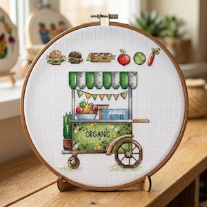 Op de afbeelding: Kruissteekborduurwerk in een houten hoepel toont een biologische voedselkar met een groen en wit luifel. De kar toont verschillende soorten fruit en groenten, en het woord "ORGANIC" is zichtbaar. Boven de kar staan afbeeldingen van een hamburger, sandwich, appel en andere voedingsmiddelen.