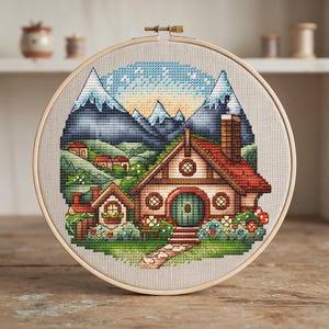 May include: A circular wooden embroidery hoop containing a cross-stitch design of a cozy cottage with a round green door, a red roof, and a stone chimney. The scene features a stone path, colorful flowers, rolling green hills, and snow-capped mountains.