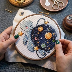 Può includere: Un telaio da ricamo a punto croce con un disegno di tartaruga celeste. La tartaruga è azzurra con un guscio blu scuro raffigurante pianeti e stelle. Il ricamo è in un telaio di legno, con stelle dorate e una spilla a forma di luna.