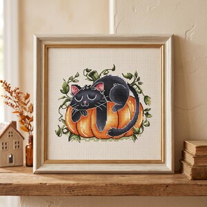 May include: A framed cross-stitch embroidery featuring a black cat with closed eyes resting on a large orange pumpkin, surrounded by green vines and leaves. The artwork is encased in a square, light-colored wooden frame with a gold inner border.