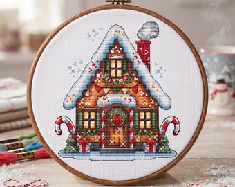 May include: A circular wooden embroidery hoop containing a cross-stitch pattern of a gingerbread house. The house features a snow-covered roof, a red chimney with smoke, candy cane accents, green shutters, and a wreath on the front door.