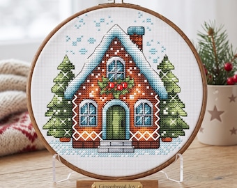 May include: A cross-stitch embroidery in a round wooden hoop featuring a gingerbread house with snow-covered roof, green pine trees, and holly accents. The house has a brown brick pattern, blue windows, and a green door. A plaque reads Gingerbread Joy.
