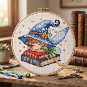 Op de afbeelding: Kruissteekborduurwerk in een houten hoepel toont een slapende fee met een blauwe tovenaars hoed met sterren. De fee rust op een stapel boeken. Het borduurwerk is omgeven door een houten hoepel, met borduurgaren en een schaar op de achtergrond.