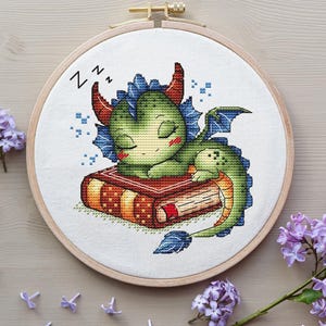 May include: Cross-stitch embroidery of a sleeping green dragon on a red book, framed in a wooden hoop. The dragon has red horns and blue accents. The word "Zz" is stitched above the dragon's head, indicating sleep. Lilac flowers are in the background.