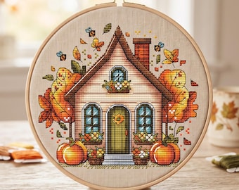 May include: A circular wooden embroidery hoop containing a cross-stitch design of a house with a green door, orange pumpkins, and autumn leaves in shades of yellow, orange, and red. Small bees are stitched flying around the roof and foliage.