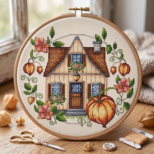 Könnte beinhalten: Ein Kreuzstich-Stickrahmen mit einer detaillierten Hausszene mit braunem Dach, blauen Fensterläden und einem Kürbis. Das Design ist von floralen und Rankenmotiven umgeben, mit einem Holzrahmen. Eine Schere und Faden sind ebenfalls sichtbar.