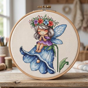 May include: A cross-stitch embroidery hoop featuring a fairy with silver hair, a purple dress, and blue wings, seated on a blue flower. The fairy wears a floral crown with red, purple, and blue flowers. The embroidery is set in a wooden hoop.