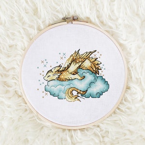 Może przedstawiać: Obręcz do haftu krzyżykowego z złotym smokiem spoczywającym na niebieskiej chmurze. Smok ma szczegółowe skrzydła i długi ogon. Tło jest białe, a obręcz wykonana jest z drewna.