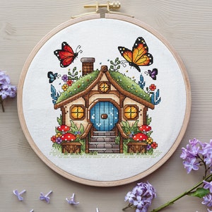 May include: A cross-stitch embroidery hoop with a whimsical illustration of a small house with a green roof, a blue door, and two butterflies. The house is surrounded by flowers and mushrooms.