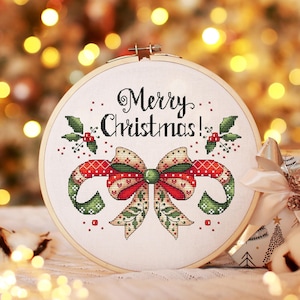 Puede incluir: Un bastidor de bordado de punto de cruz con temática navideña. El diseño presenta un lazo y bastones de caramelo rojos y verdes, con las palabras "Merry Christmas!" en escritura negra. El bastidor se encuentra sobre un fondo borroso de luces navideñas.