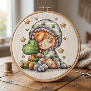 Puede incluir: Un bastidor de bordado de punto de cruz con un niño con un traje con capucha de lunares verdes, abrazando a un dinosaurio verde. El diseño incluye estrellas doradas y está sobre tela crema, en un aro de madera.