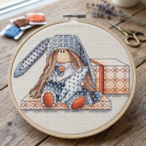 May include: Cross-stitch embroidery of a bunny wearing a blue hat and holding a star-shaped object. The bunny is sitting on a patterned surface, with a gift box beside it. The embroidery is set in a wooden hoop.