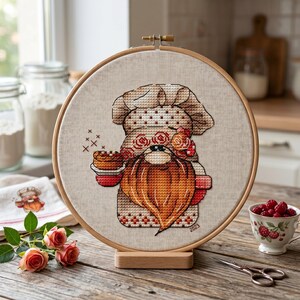 May include: A cross-stitch embroidery of a gnome baker wearing a chef's hat and holding a cupcake. The gnome has a long orange beard and is surrounded by roses. The embroidery is set in a wooden hoop and displayed on a wooden stand.