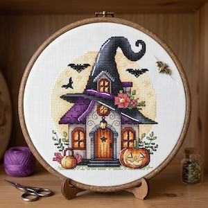 May include: A cross-stitch embroidery of a Halloween witch's house, featuring a black witch hat, purple roof, and a jack-o'-lantern. The design is set in a wooden embroidery hoop, with a wooden stand.