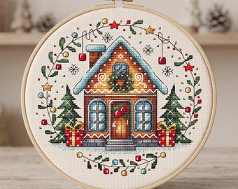 May include: A circular wooden embroidery hoop containing a cross-stitch pattern of a gingerbread house with a snow-covered roof, a wreath on the door, two green pine trees, and two red gift boxes, all framed by a decorative holiday garland.