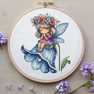 May include: A cross-stitch embroidery of a fairy with a flower crown sitting on a blue bell flower. The fairy has a purple dress and wings. The background is white.