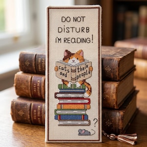 May include: A rectangular cross-stitch bookmark on cream fabric with a brown border and tassel. It features the text DO NOT DISTURB I'M READING! above a calico cat holding a book titled cats and their people, perched on a stack of colorful books.