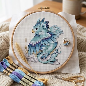 May include: Cross-stitch embroidery of a blue and teal griffin with a golden beak and claws, set in a wooden hoop. The design includes wheat stalks. Embroidery floss in shades of blue, purple, and gold, along with gold scissors and a needle minder are also visible.