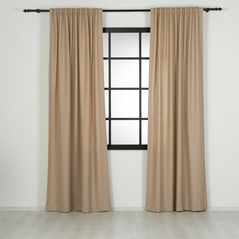 May include: A pair of beige curtains hanging in a window with a black window frame. The curtains are made of a soft, textured fabric.