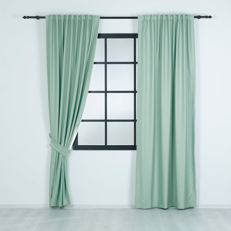 May include: A pair of light green curtains hanging on a window with a black frame. The curtains are drawn back on one side, revealing the window behind.