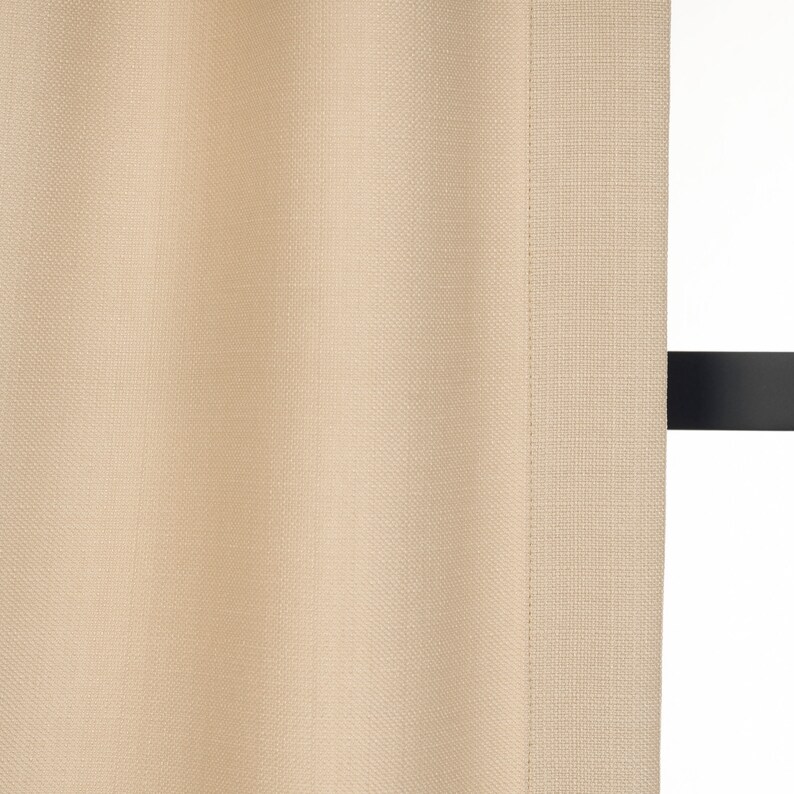 May include: A close-up of a beige curtain with a subtle textured weave. The curtain is hanging from a black rod.