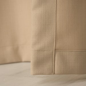 May include: Close-up of the bottom edge of a beige curtain panel. The fabric has a woven texture and a subtle sheen.