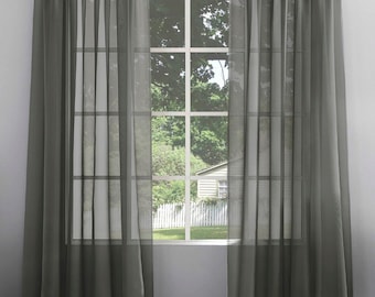 May include: A pair of sheer grey curtains hang in front of a window with a view of a house and trees. The curtains are made of a lightweight fabric that allows light to filter through.