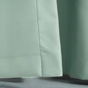 May include: Close-up of a light green curtain panel with a stitched hem and a decorative seam running down the side.