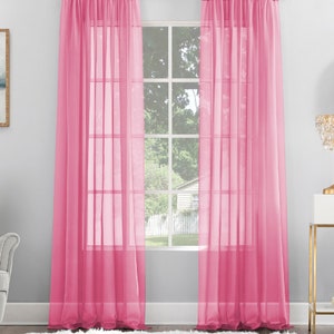 May include: Two sheer pink curtains hang in front of a window with a view of a house and trees. The curtains are made of a lightweight fabric that allows light to filter through.