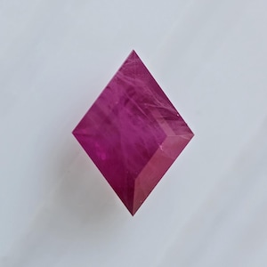 May include: A pink, faceted, diamond-shaped crystal with a translucent appearance.