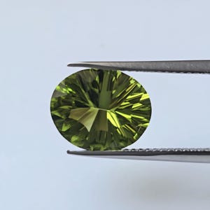 May include: A round, faceted, olive-green gemstone is held by a pair of silver tweezers against a white background. The gemstone's cut creates a starburst pattern, reflecting light. The gem is a vibrant, translucent green.