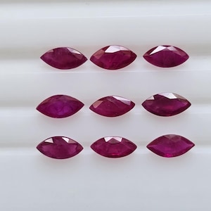 Puede incluir: Nueve rubíes de talla marquesa, cada uno con un tono rojo intenso, están dispuestos en tres filas sobre una superficie blanca. Las piedras preciosas tienen un diseño facetado, que capta la luz y muestra sus superficies pulidas. Estos rubíes son ideales para la joyería.