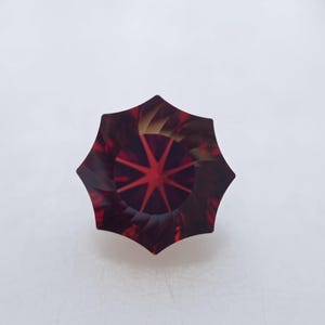 May include: A deep red gemstone with a star-shaped pattern cut into the center. The gemstone is faceted and has a hexagonal shape.