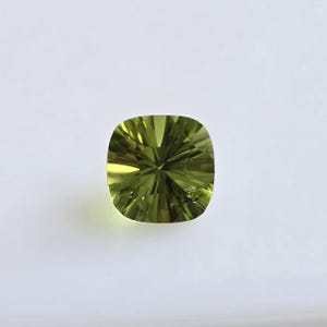May include: A faceted, cushion-cut gemstone in shades of olive green. The gem has a symmetrical pattern of facets that reflect light, creating a sparkling effect. The gemstone is set against a plain white background.